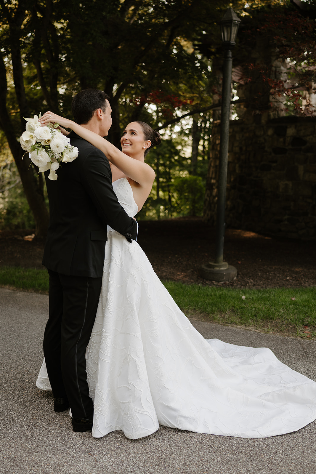 Bride smiling up at the groom while embracing on a paved path next to a vintage lamppost.
